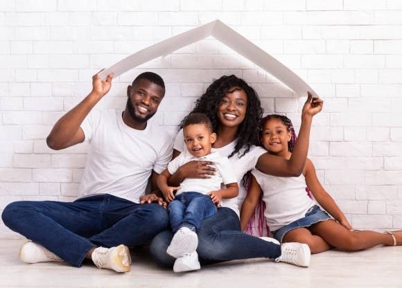 Family Housing Concept. Young African American Parents With Two Children Sitting Under Cartboard Roof Dreaming Of New Home, Copy Space